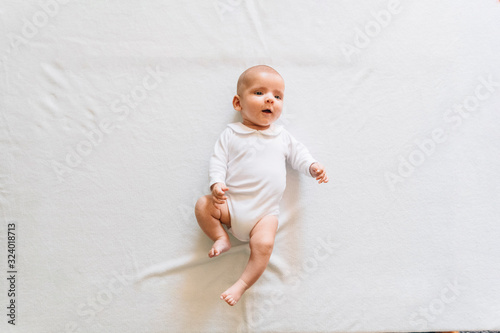Top view of lovely infant in white pajama with open mouth lying on bed moving arms and legs looking up