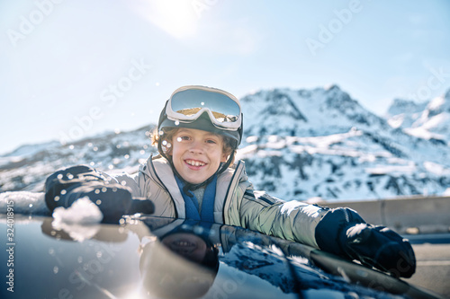 Delighted boy in helmet with goggles smiling and rolling snowball while peeking out vehicle window on sunny day on ski resort photo