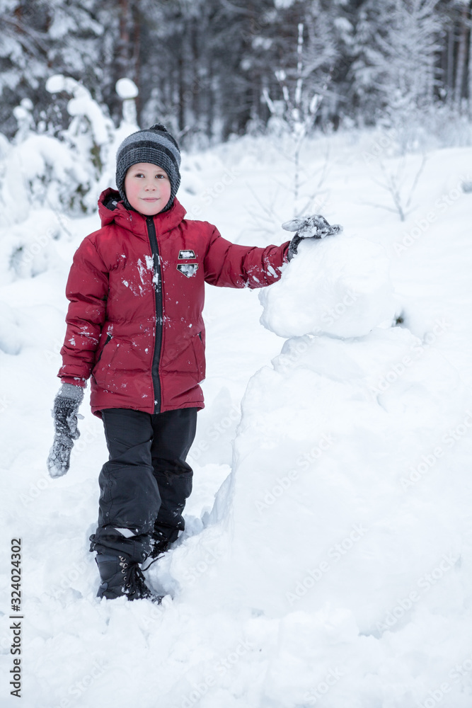 Caucasian boy in red jacket standing near snowman in forest