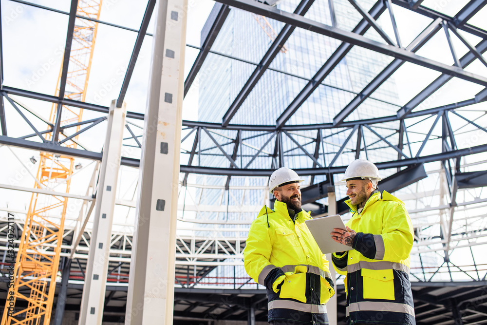 Men engineers standing outdoors on construction site, using tablet.