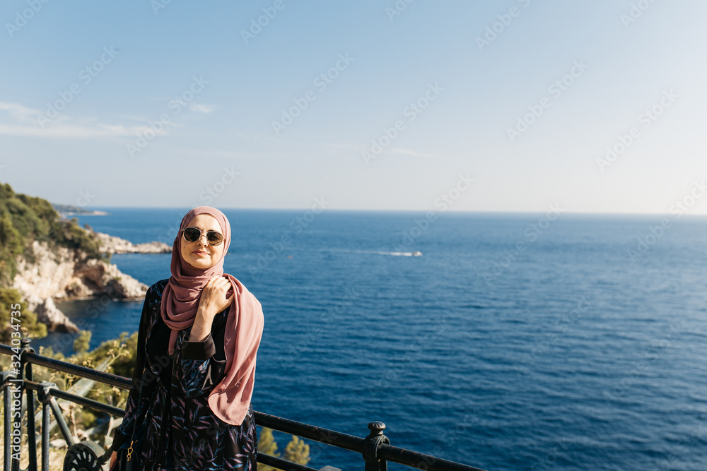 portrait of young European Muslim women with hijab standing and looking at the camera. Sea is in the background. She is happy and relaxed.	