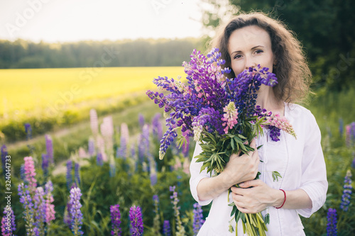 Beautiful woman in a white dress holds a large bouquet of lupins in the field.