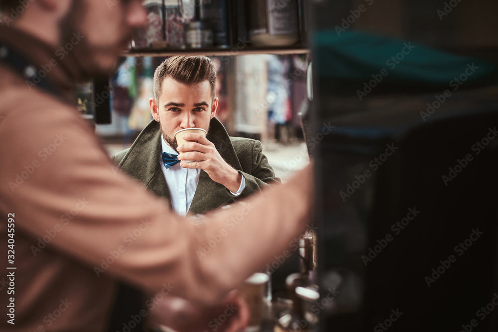 Stylish and elegant young male customer sitting outdoors next to coffee making barista in a mobile coffee shop in a city emporium, wearing green wool coat, white shirt and bow tie, taking a sip of Stylish and elegant young male customer sitting outdoors next to coffee making barista in a mobile coffee shop in a city emporium, wearing green wool coat, white shirt and bow tie, taking a sip of