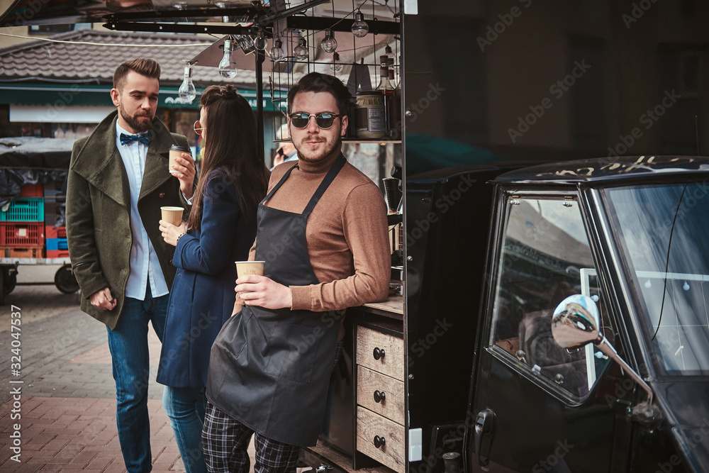 Charming and calm male barista standing relaxed outdoors near mobile coffee shop and two elegant looking, chatting customers wearing dark apron, sunglasses and warm wool brown sweater in a combination