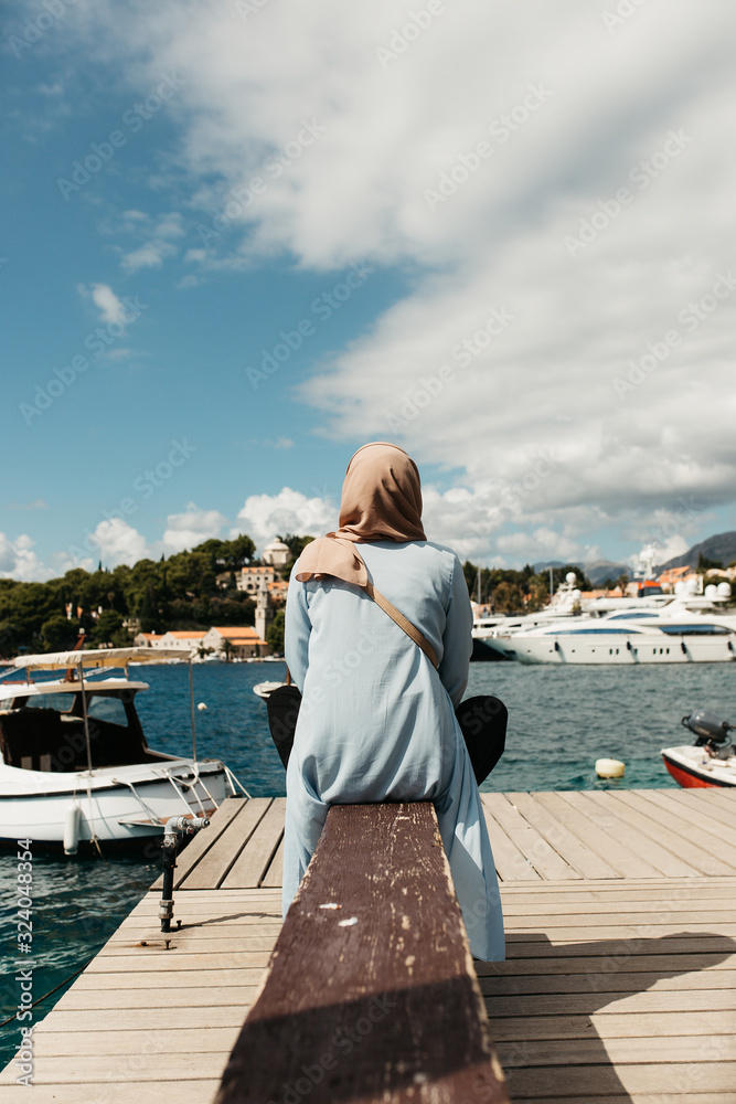 woman with hijab sitting on deck by the sea.  Female tourist exploring Cavtat. She is sitting turned back and looking in the old city. 