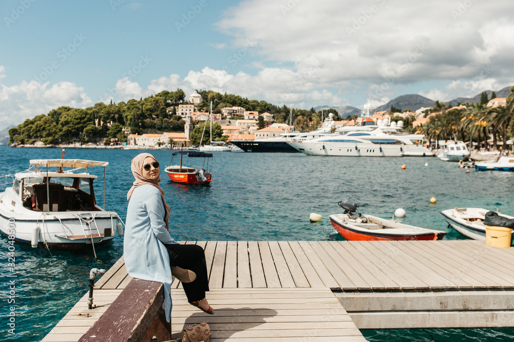 woman with hijab sitting on bench by the sea.  Female tourist exploring Cavtat. She is looking to the camera. 