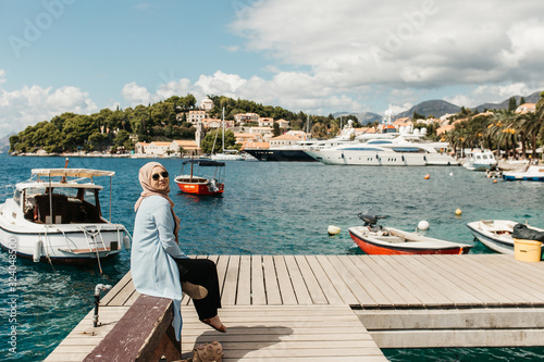 woman with hijab sitting on bench by the sea.  Female tourist exploring Cavtat. She is looking to the camera.  © Melika