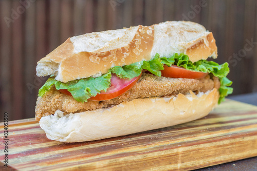 Tasty big fried chicken sandwich on french bread with tomatoes and lettuce on wooden table. Top view.