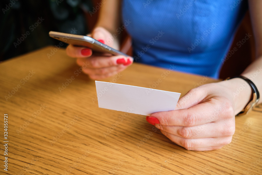 A girl in a blue dress sits at a cafe and writes a new contact on the phone with a business card.