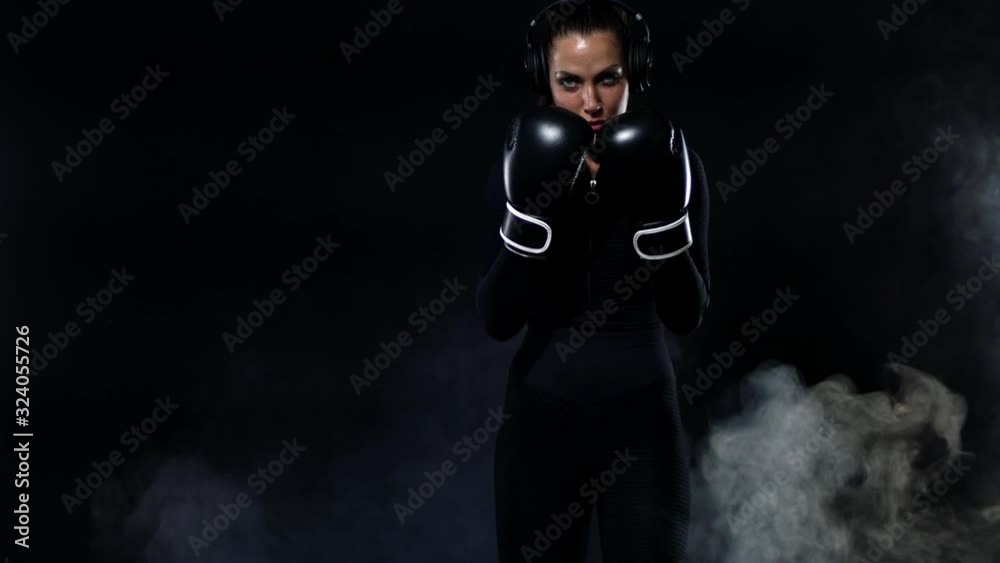 Young woman sportsman boxer doing boxing training at the gym. Girl wearing gloves, sportswear and hitting the punching bag. Isolated on black background with smoke. Copy Space. Young woman sportsman boxer doing boxing training at the gym. Girl wearing gloves, sportswear and hitting the punching bag. Isolated on black background with smoke. Copy Space.