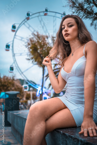 Street style, portrait of a beautiful young brunette in white dress in front of the tourist ferris wheel in the city