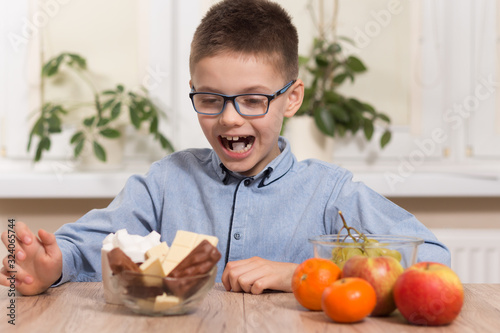 Chłopiec w okularach siedzi przy stole i z zadowoloną miną wpatruje się w słodycze. Obok leżą owoce mandarynki, jabłka i winogron.