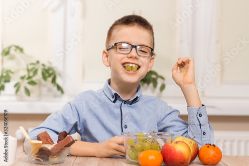 Uśmiechnięty chłopiec siedzi przy stole i zjada owoce. Trzyma w zębach owoce winogrona.