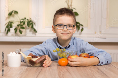 Uśmiechnięty chłopiec w wieku szkolnym siedzi przy stole i przysuwa do siebie owoce. Drugą ręką odsuwa od siebie naczynia ze słodyczami.
