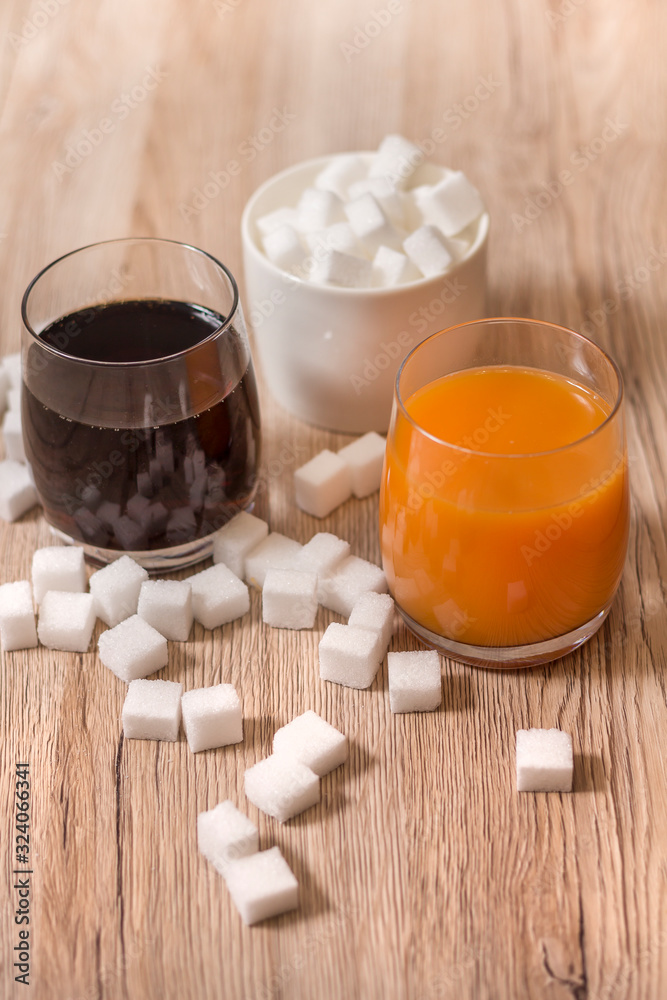 Kostki cukru porozrzucane leżą na blacie kuchennym. W jednej szklance stoi cola a w drugiej sok. Na drugim planie cukiernica wypełniona kostkami cukru.