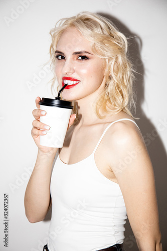 smiling young beautiful blonde girl in a white T-shirt drinks from a disposable cup through a straw on a white background, studio fashion photo