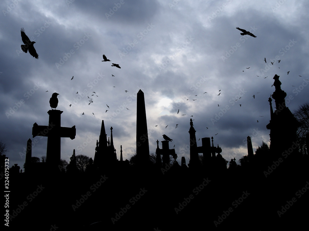 crows flying and perched on old gothic style gravestone in silhouette with tall memorials and crosses against an overcast cloudy sky