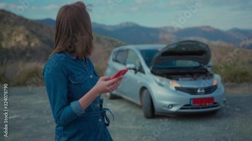 Young upset woman standing at broken car searching for repair service on smartphone worrying in stress. Car accident. Car trouble.