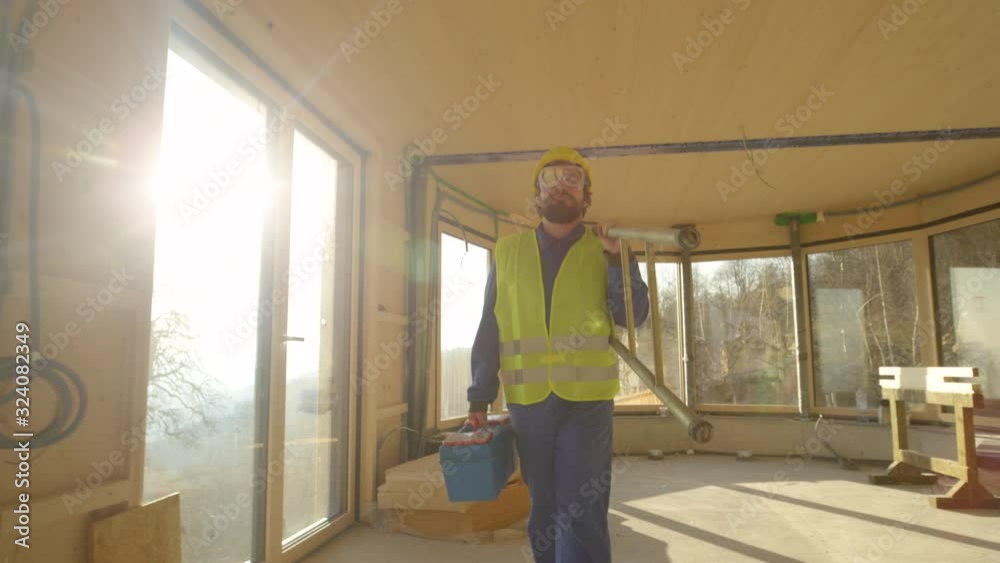 SLOW MOTION LENS FLARE CLOSE UP: Content builder walks out of a CLT house after a long day at work. Caucasian contractor carries his toolbox and ladder out of a prefabricated house under construction. SLOW MOTION LENS FLARE CLOSE UP: Content builder walks out of a CLT house after a long day at work. Caucasian contractor carries his toolbox and ladder out of a prefabricated house under construction.