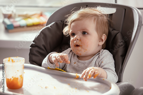  Baby in the child dining chair eat healthy  vegetable food. Child eat  pumpkin and carrot puree with banana. Sunny day. Dirty face of happy kid. Portrait of a baby eating with a stained face.