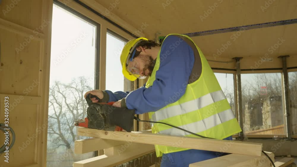 SLOW MOTION, LENS FLARE, CLOSE UP, DOF: Young Caucasian man working on a prefabricated house buffs a plank with a sander. Builder wearing a helmet and yellow vest sands a wooden beam on a sunny day. SLOW MOTION, LENS FLARE, CLOSE UP, DOF: Young Caucasian man working on a prefabricated house buffs a plank with a sander. Builder wearing a helmet and yellow vest sands a wooden beam on a sunny day.