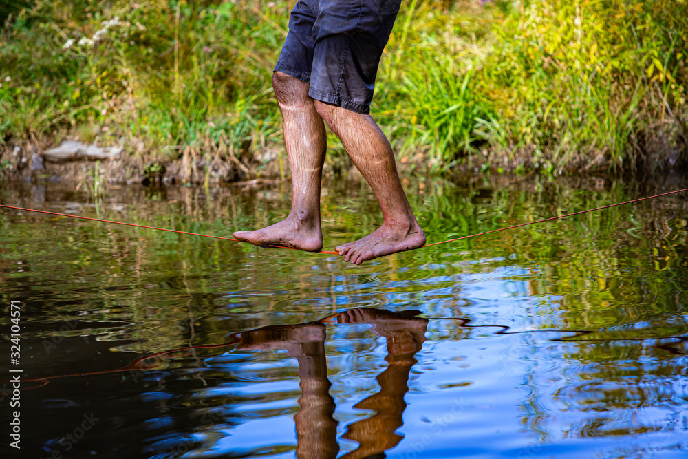 A healthy man wearing shorts with soaked legs is seen balancing on a slackline over a pond in nature, barefooted and close to surface with reflection A healthy man wearing shorts with soaked legs is seen balancing on a slackline over a pond in nature, barefooted and close to surface with reflection