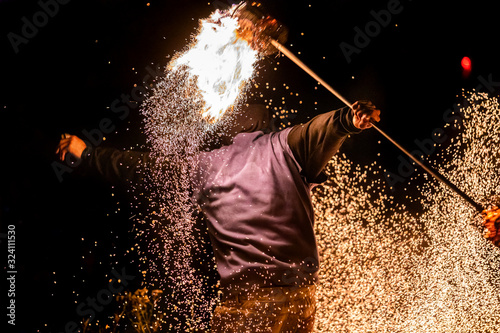 Canvas-taulu A close up energetic shot of a fire dance performer using a long flaming pole to create flying embers and light effect