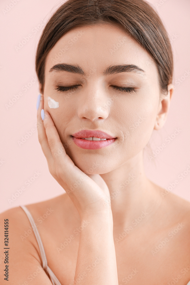 Beautiful young woman applying cream on her face against color background