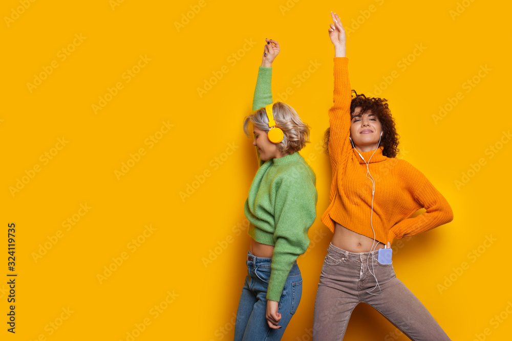 Obraz premium Two gorgeous caucasian girls dancing on a yellow background while listening to music through headphones
