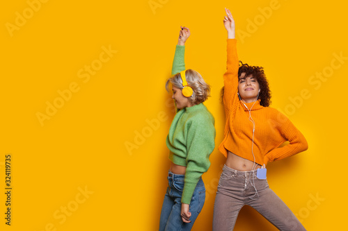 Two gorgeous caucasian girls dancing on a yellow background while listening to music through headphones