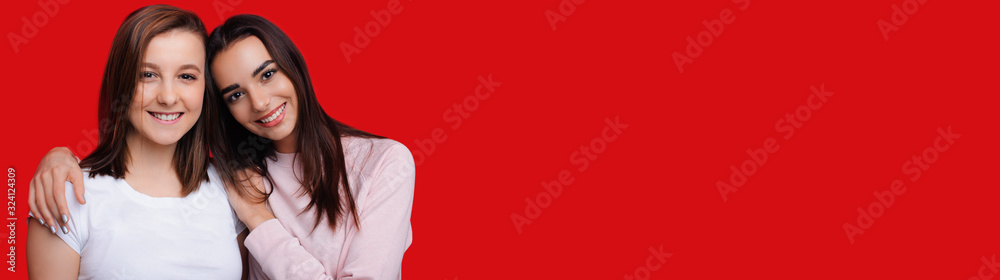 Two lovely caucasian girls embracing and smiling are posing on red background with blank space Two lovely caucasian girls embracing and smiling are posing on red background with blank space