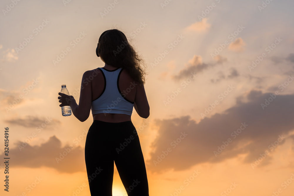 Silhouette young woman in the headphones in the sport clothes with bottle of clear mineral water on the sunset sky backgrounds. The concept of healthy lifestyle