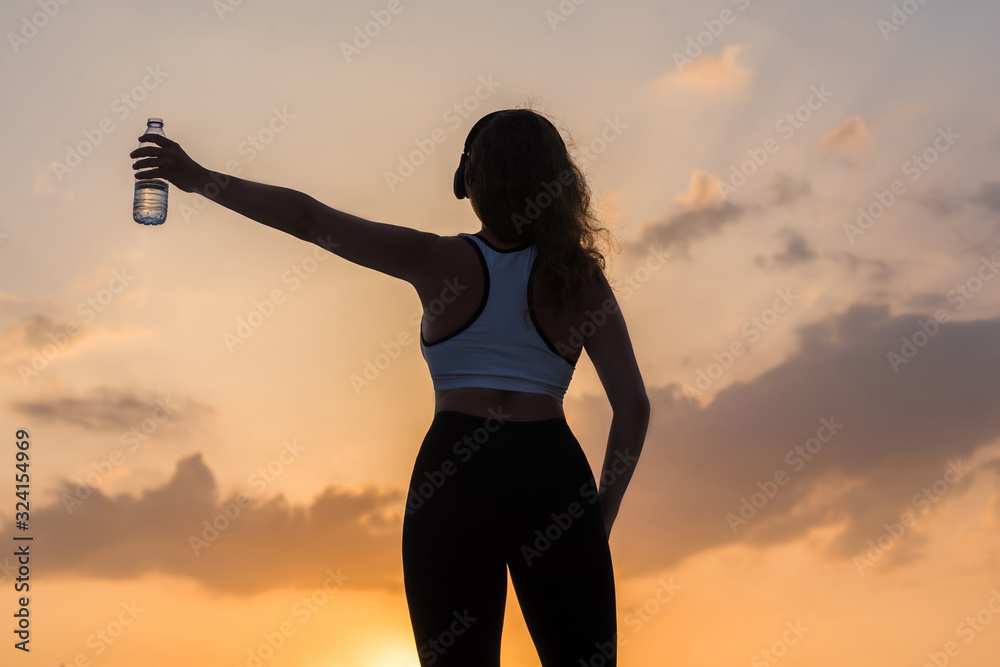 Silhouette of happy young woman in the sport clothes with bottle of clear mineral water on the sunset sky backgrounds. The concept of healthy lifestyle