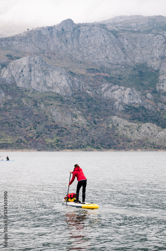 A girl in a red coat practices paddle surfing exploring on a mountain lake with luggage on top of her board