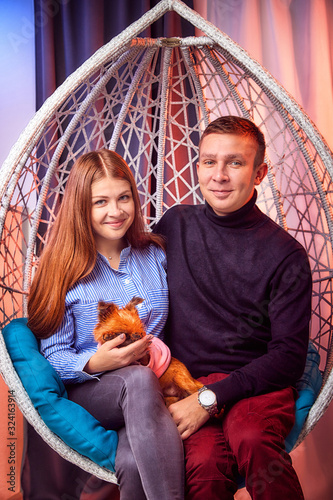 Beautiful brunette girl whth long hair in the blue shirt and man in black sweater and small dog on a white wicker hanging chair with pillow photo