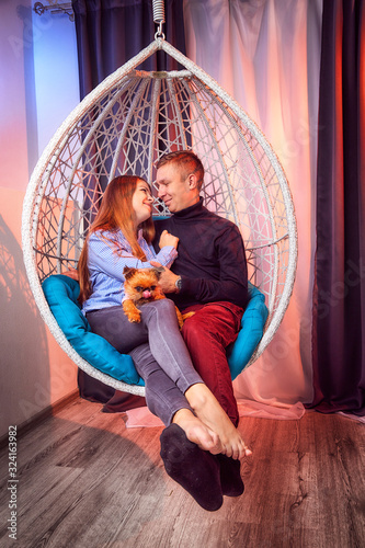 Beautiful brunette girl whth long hair in the blue shirt and man in black sweater and small dog on a white wicker hanging chair with pillow photo