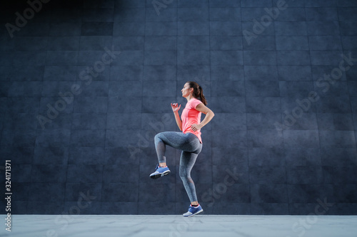 Side view of fit attractive Caucasian woman in sportswear and with ponytail running outdoors. In background is dark wall. © dusanpetkovic1