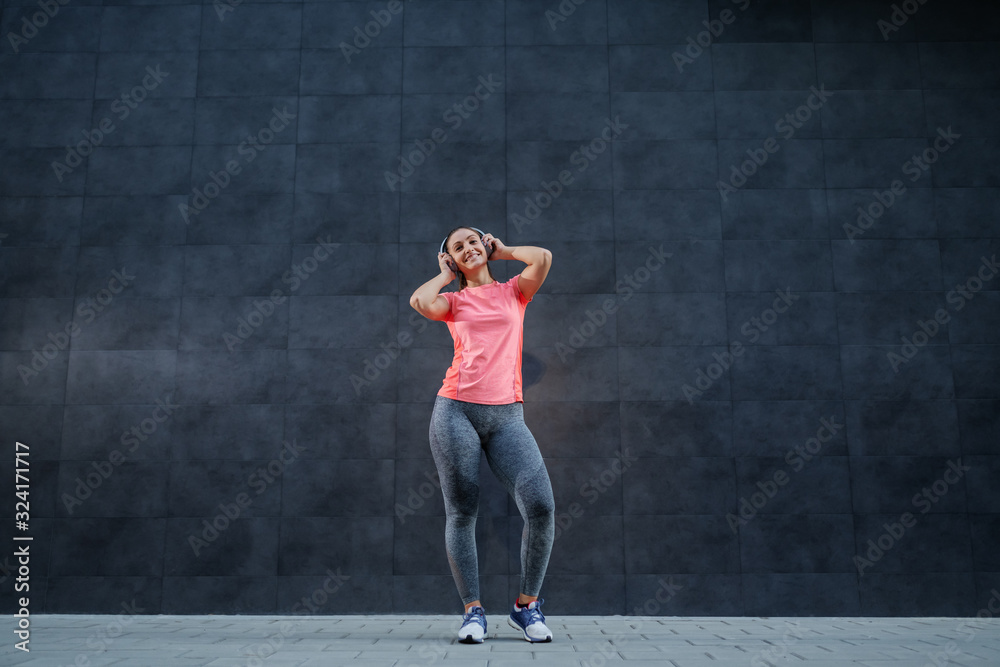 Full length of attractive smiling Caucasian brunette in sportswear and with ponytail listening music over headphones. In background is dark wall. Full length of attractive smiling Caucasian brunette in sportswear and with ponytail listening music over headphones. In background is dark wall.