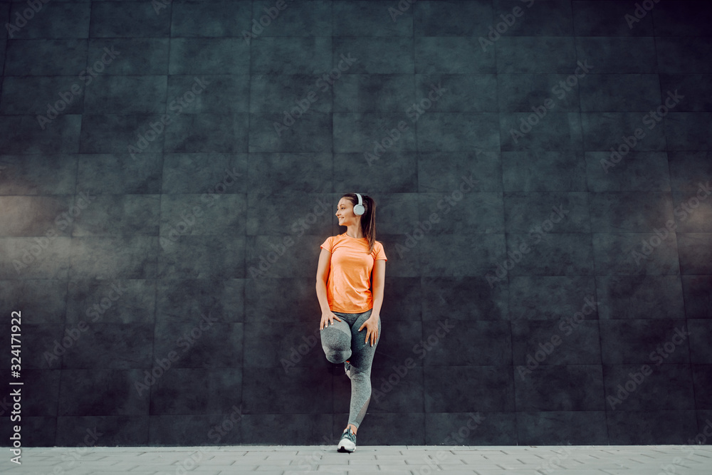 Full length of attractive smiling Caucasian brunette in sportswear and with ponytail listening music over headphones while leaning on the wall. In background is dark wall.