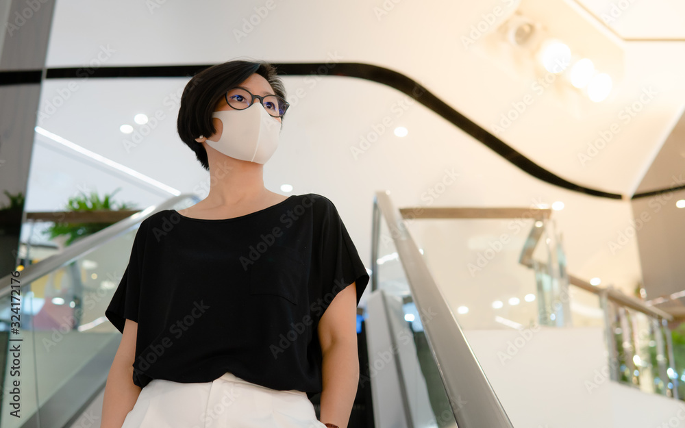 Beautiful middle aged Asian woman stand on escalator, wear medical face mask to protect from infection of virus, pandemic, influenza outbreak and epidemic of disease in empty shopping mall. Beautiful middle aged Asian woman stand on escalator, wear medical face mask to protect from infection of virus, pandemic, influenza outbreak and epidemic of disease in empty shopping mall.