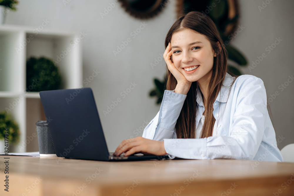 Young modern girl office worker sits at a table with a laptop in the office and smiling Young modern girl office worker sits at a table with a laptop in the office and smiling