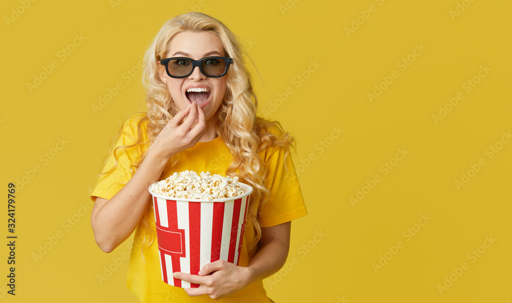 Surprised young blond woman in 3d glasses and yellow shirt eating popcorn, looks shocking movie at the cinema. Isolated on yellow background
