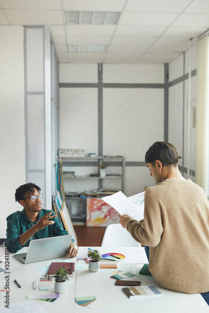 Two young designer sitting at the table and discussing sketches of their new project in workshop Two young designer sitting at the table and discussing sketches of their new project in workshop