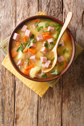 Spicy hot pea soup with potatoes, onions, carrots and ham close-up in a bowl. Vertical top view