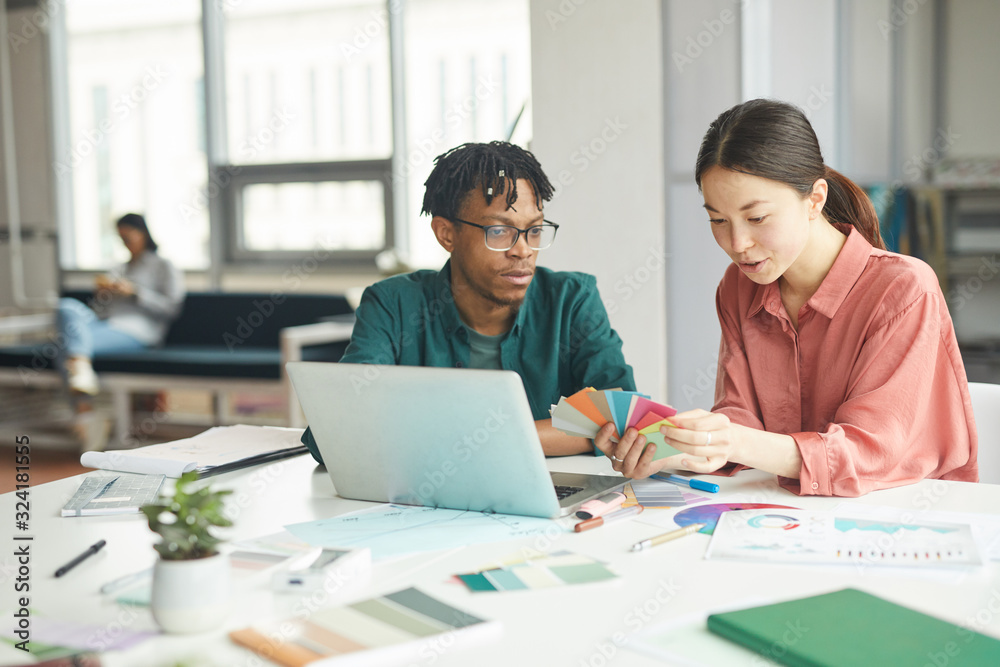 African young designer sitting at the table in front of laptop with his colleague and choosing color for their new project while they working African young designer sitting at the table in front of laptop with his colleague and choosing color for their new project while they working