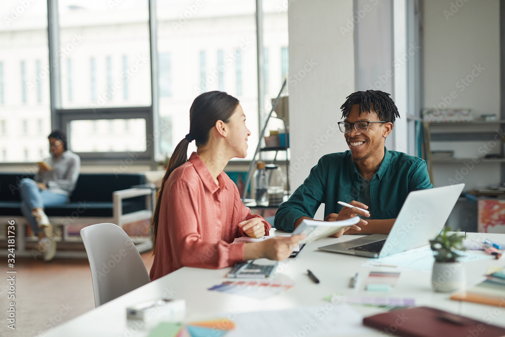 Young multiethnic business couple smiling and talkign to each other while sitting at the table with laptop at office Young multiethnic business couple smiling and talkign to each other while sitting at the table with laptop at office
