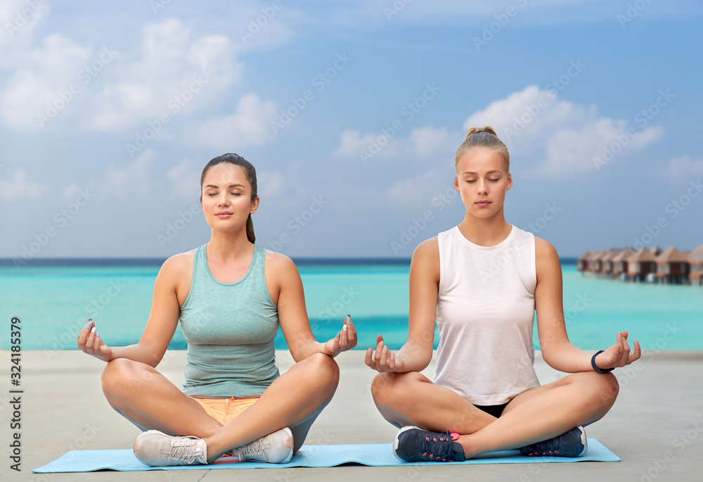 fitness, yoga and healthy lifestyle concept - young women or female friends meditating in lotus pose on mat over sea and bungalow on background