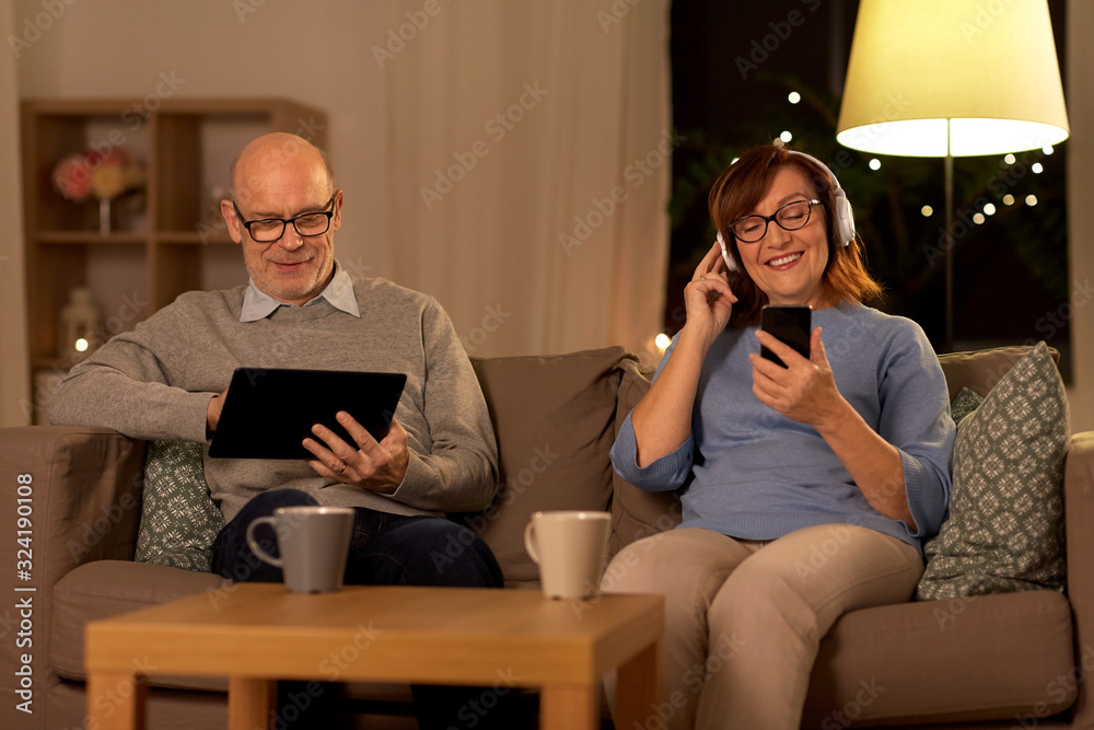 technology, old age and people concept - happy senior couple with tablet pc computer, headphones and smartphone at home in evening technology, old age and people concept - happy senior couple with tablet pc computer, headphones and smartphone at home in evening