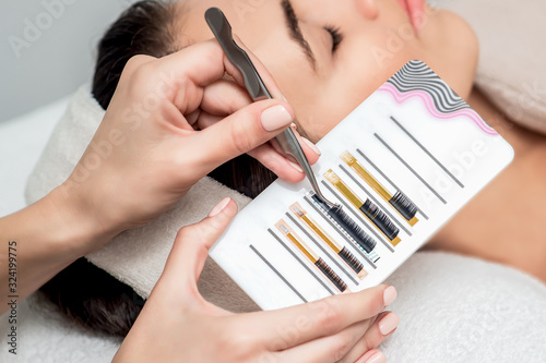 Hands of beautician holding artificial eyelashes by tweezers next to face of young woman, close up.