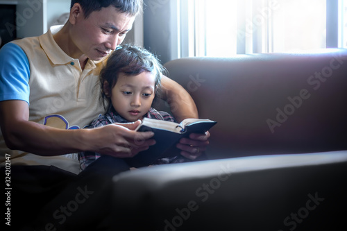 Father reading bible to daughter on sofa in morning at home, family christian concept.